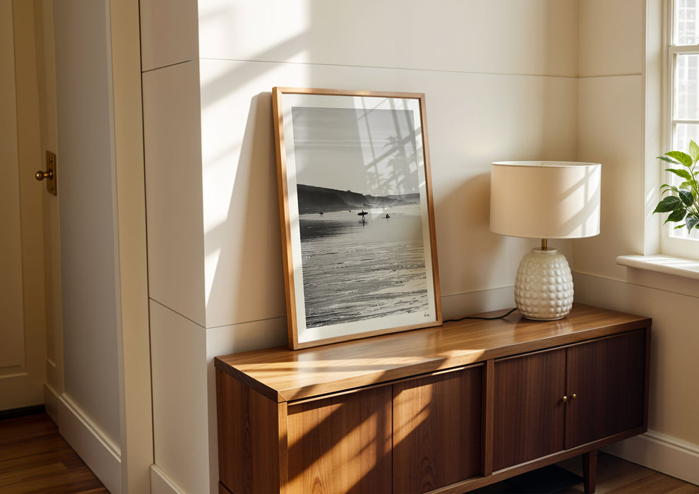 Clear To Land, a black-and-white photo print of surfers on the beach at Polzeath, Cornwall in a frame against a wall.