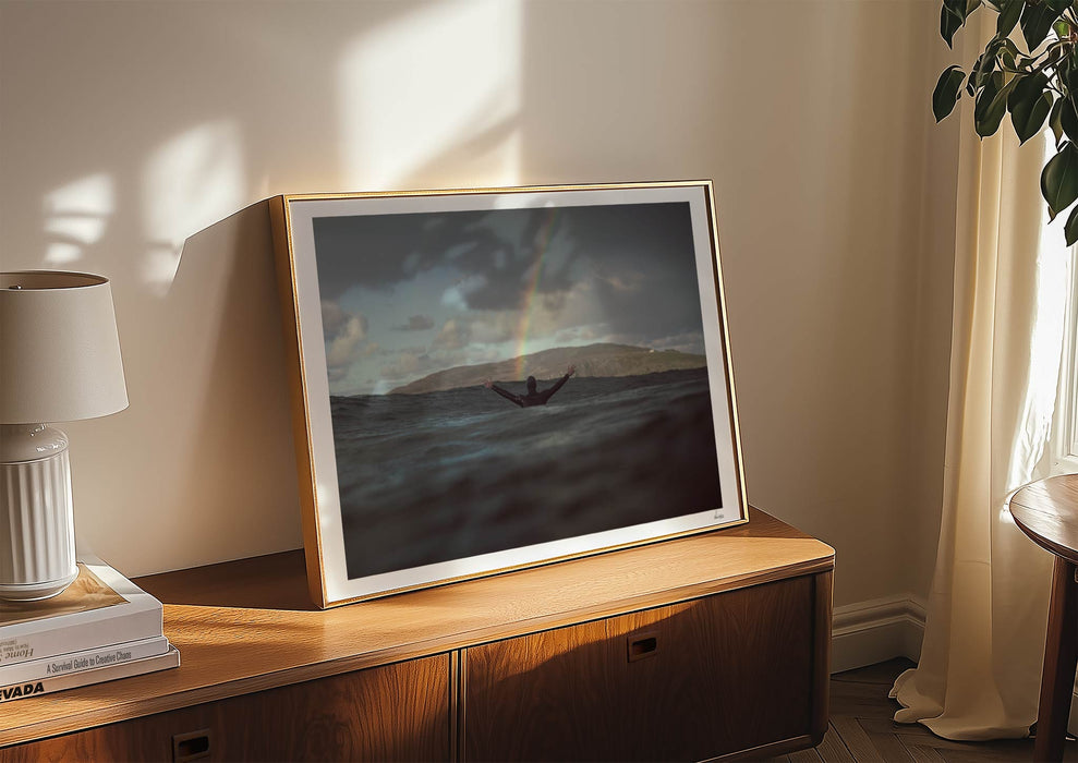 A framed print in a room of Colours With Colour, a colour photograph of a surfer saluting a rainbow in Scotland.