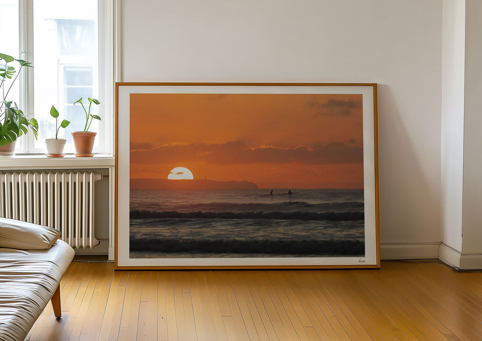 Watching Her Sink, a colour photographic print of surfers at sunset at Croyde Bay, Devon in a frame leaning against a wall.