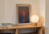 We’ll Meet Again, a colour photographic print of a bird and sunset over a calm ocean in a wooden frame on a shelf.